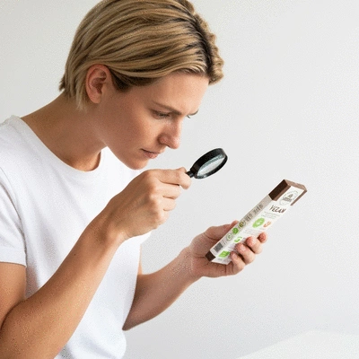 Person thoughtfully examining a vegan chocolate bar with a magnifying glass, focusing on the ingredient list and certifications, clean background, natural lighting, no text, no words, no typography, no labels, clean image